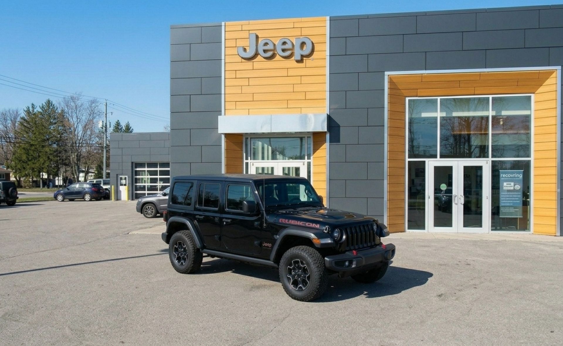 A black four-door Jeep Wrangler Rubicon SUV parked on the paved lot in front of a modern Jeep dealership showroom building with distinct dark grey tile and light wood-paneled accents on a sunny day in Raleigh.