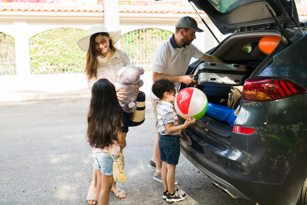 Happy beautiful family putting suitcases, floaters and a beach ball on the car. Latin parents talking with their son and daughter about summer vacations