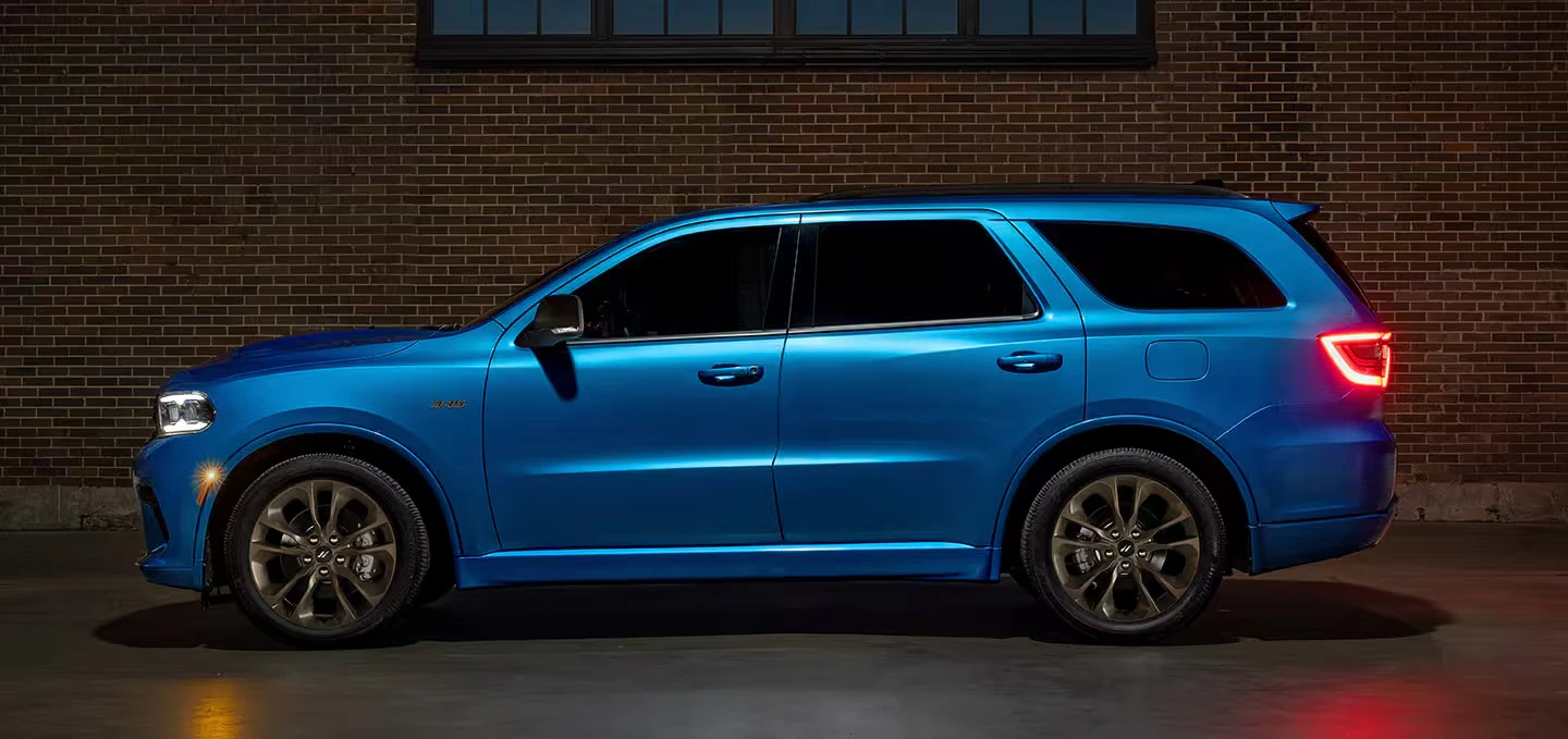 Blue 2027 Dodge Durango SUV in side profile with bronze alloy wheels and glowing red taillights, shown in front of a dark brick wall at night