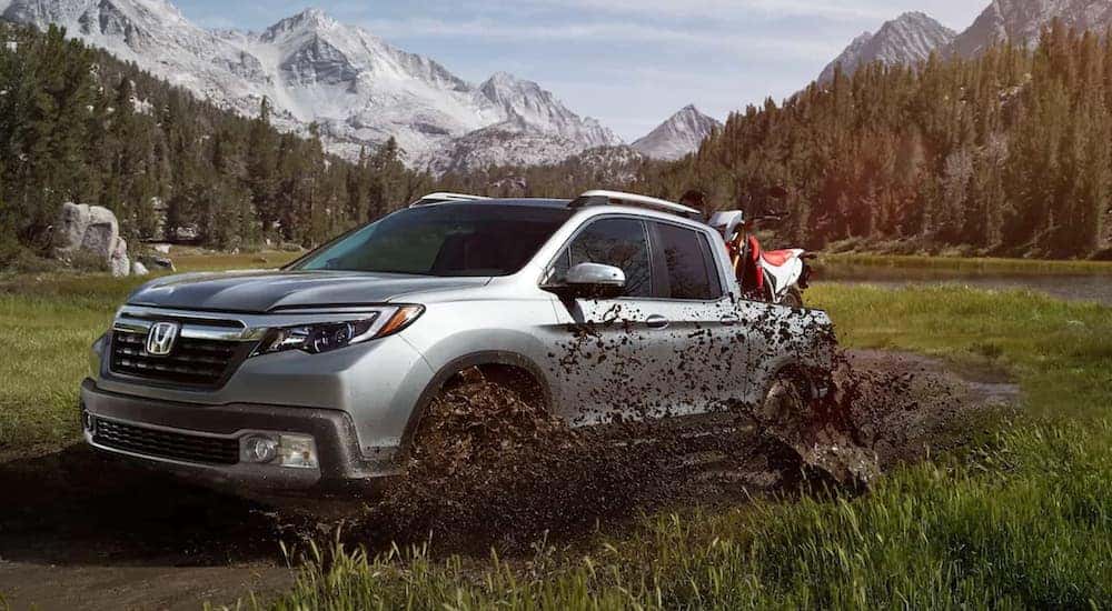 A silver 2020 Honda Ridgeline RTL-E is off-roading in the mud with a dirt bike in the bed and mountains in the distance.