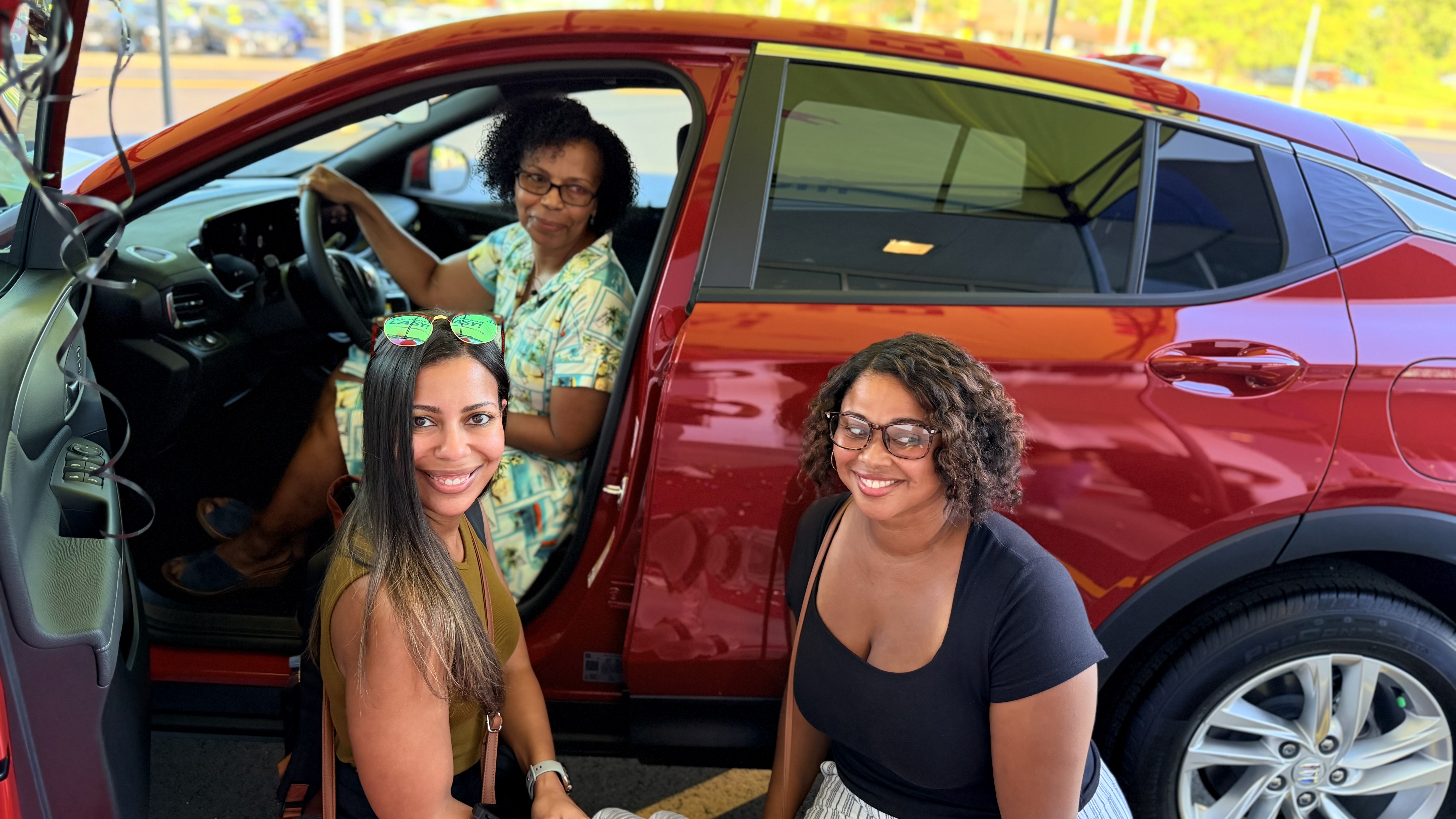 Three smiling women posing with a shiny red SUV, with one sitting in the driver's seat and two kneeling alongside the open car doors outside.