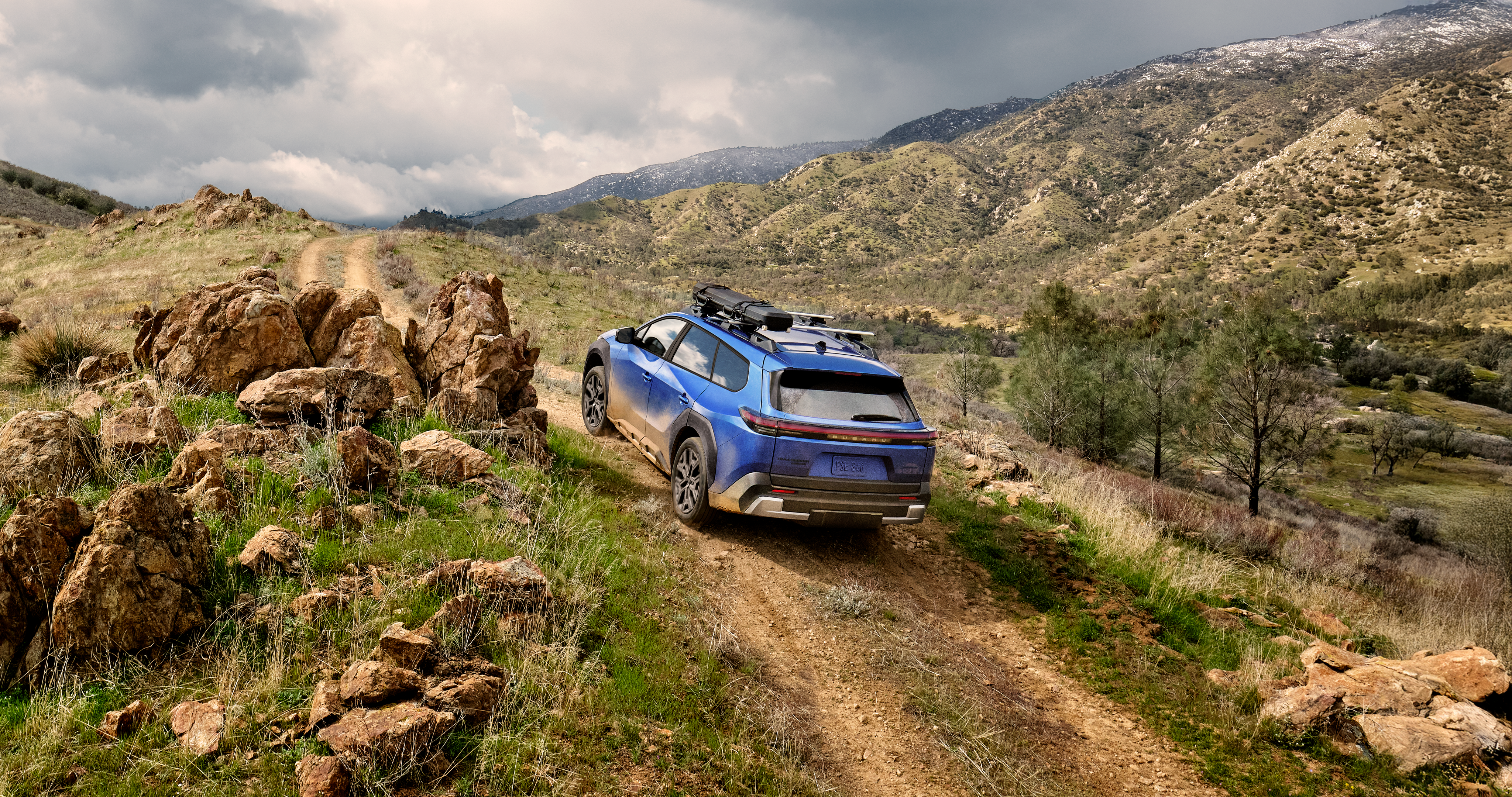 A static image of a blue 2026 Subaru Trailseeker driving on a narrow dirt trail that winds through rocky, grassy hills. 