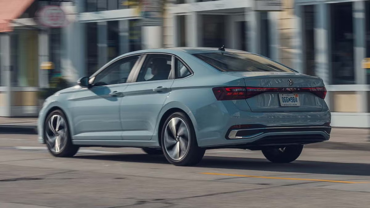 2027 Volkswagen Jetta in light blue driving on a city street, rear three-quarter view
