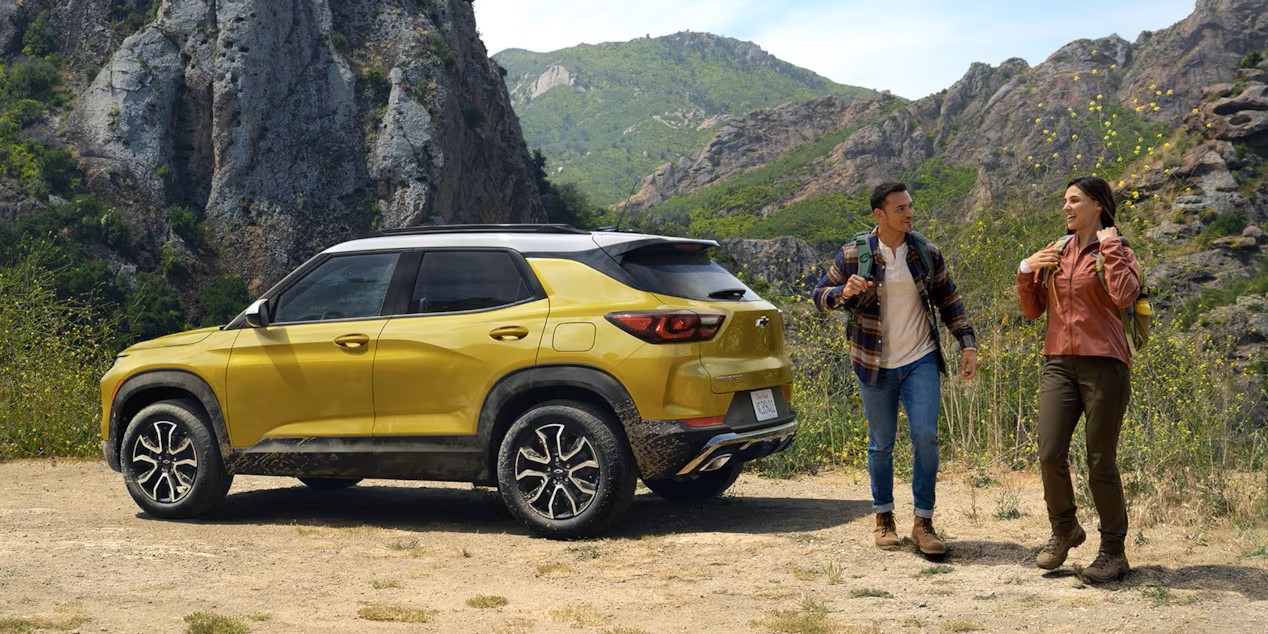 a yellow compact suv parked on a dirt road in the mountains with 2 hikers next to it