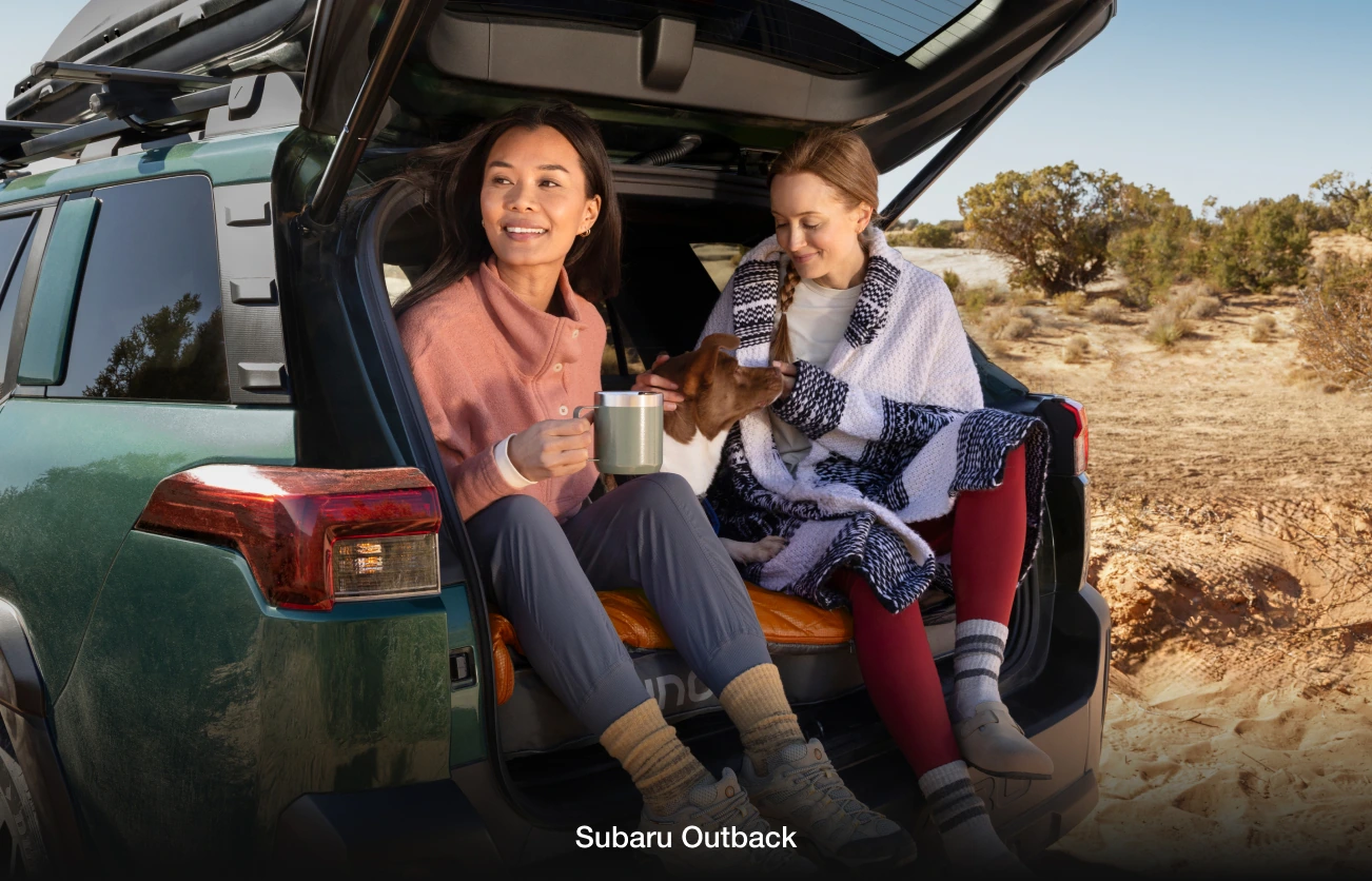 Two women sit in the back of a green Subaru Outback, enjoying a desert scene.