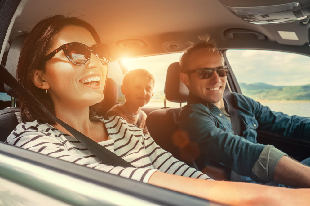 Two parents sit in the front of the car, both with sunglasses on and smiling with their kid in the back seat leaned forward and smiling, there is sun in the background and they're driving through hilly terrain