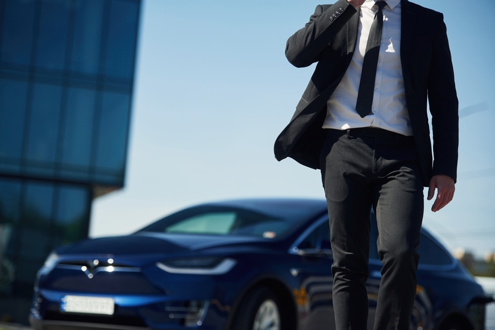 man in business suit pictured from the shoulders down, walking away from his car that's parked outside of a glass building